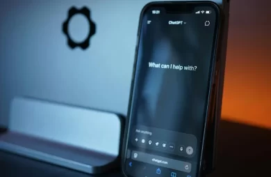 Interfaz de ChatGPT en un teléfono móvil mostrando la pantalla de inicio, con un ordenador al fondo y luz ambiental cálida.