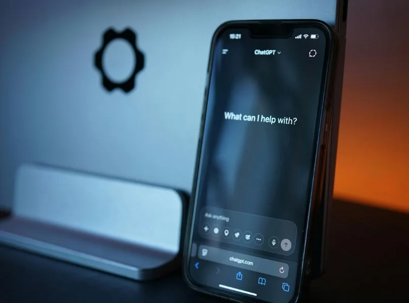 Interfaz de ChatGPT en un teléfono móvil mostrando la pantalla de inicio, con un ordenador al fondo y luz ambiental cálida.